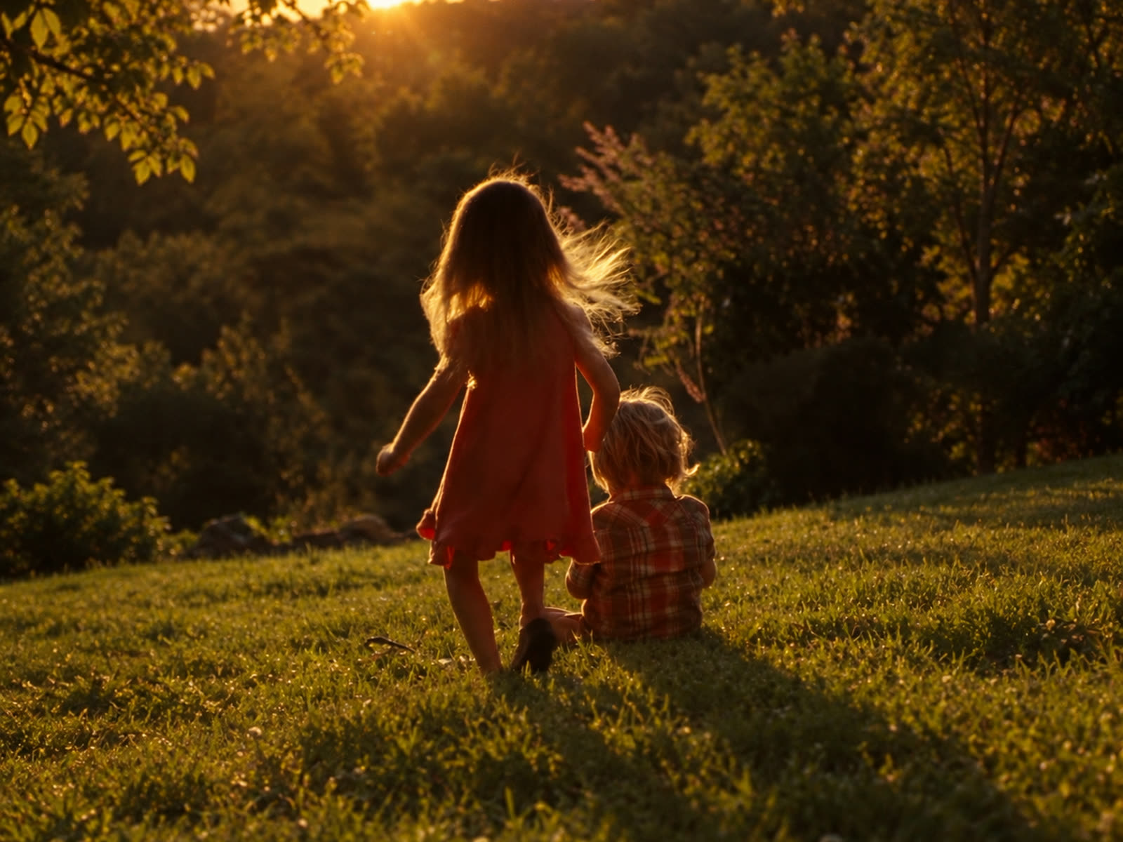 Two children in a grassy field at sunset, one standing and one sitting, watching the evening light through the trees